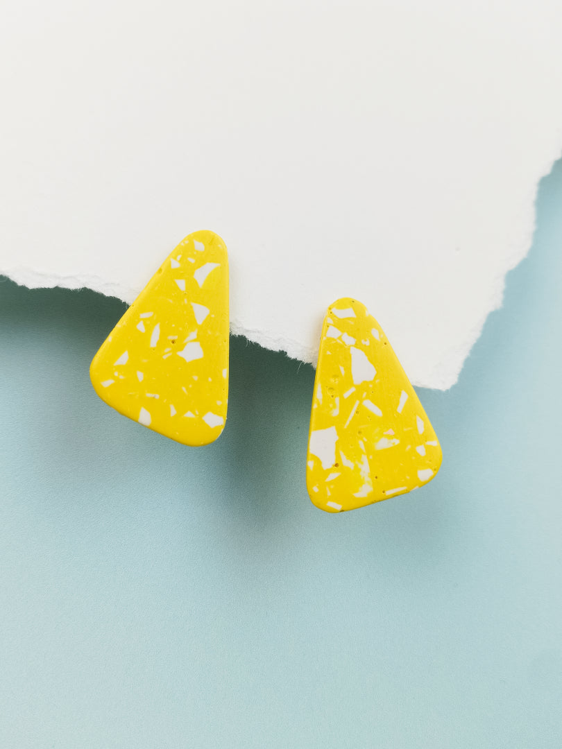 boucles d'oreilles modèle alma géométriques couleur jaune terrazzo blanc présentées de face suspendues sur un papier blanc et u fond bleu clair